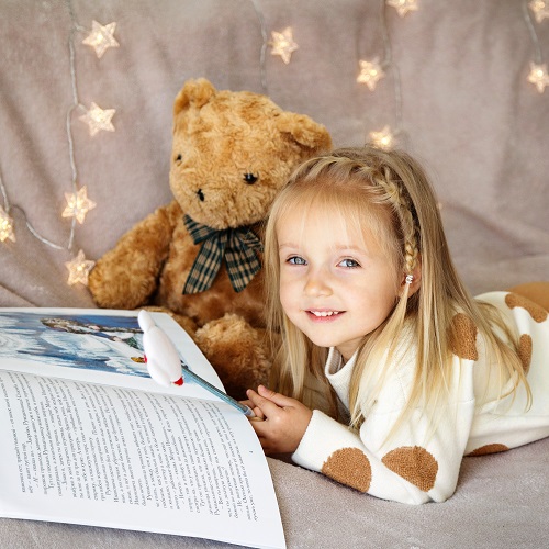 little girl lying on the couch at home and reading a children's winter wonderland, teddy bear sits next